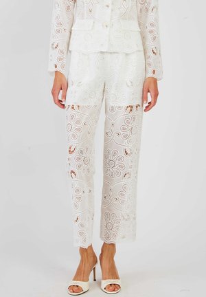 Białe, kwiatowe koronkowe spodnie z szerokimi nogawkami, noszone z dopasowaną kurtką i białymi sandałami na obcasie z odkrytymi palcami, pokazane od połowy tułowia w dół.