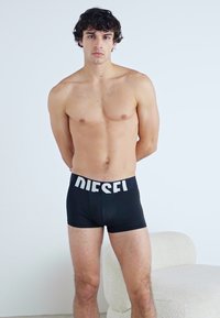 Fekete boxer alsónadrág fehér "DIESEL" logóval a derékrészen, testhezálló kialakítással, pamut anyagból, egy álló férfi viseli egy sima háttér előtt.