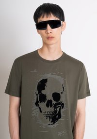 Maslinasto zelena majica s crnim grafičkim prikazom lubanje koja sadrži digitalne uzorke. Model nosi upadljive crne sunčane naočale, ističući dizajn.