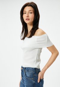 Weißes geripptes Oberteil mit schulterfreiem Schnitt und kurzen Ärmeln, kombiniert mit hochgeschnittenen blauen Jeans. Das Oberteil hat eine enganliegende Passform.