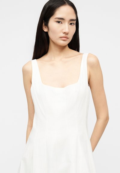 Γυναίκα με μακριά σκουρόχρωμα μαλλιά, φορώντας λευκό φόρεμα χωρίς μανίκια με τετράγωνη λαιμόκοψη, κοιτάζει ευθεία μπροστά με ουδέτερη έκφραση.