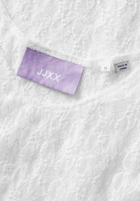 Top de encaje blanco con una textura suave, con una etiqueta lila que dice "JJXX" y tamaño "S", junto a una pequeña etiqueta de cuidado que indica "HECHO EN CHINA".