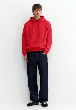 Jeune personne debout contre un mur blanc, portant un sweat à capuche rouge, un jean large foncé et des baskets noires avec des lacets blancs, une main dans la poche.