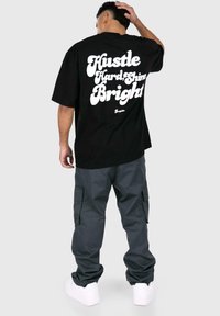 Sort bomulds t-shirt med oversized pasform, prydet med hvidt fed skrift "Hustle Hard, Shine Bright" på ryggen. Samlet med mørkegrå cargo bukser.