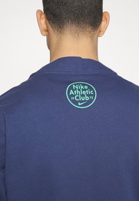 Pullover navy blu con un logo stampato verde a forma rotonda che recita 'Nike Athletic Club 1972' nella parte superiore della schiena. Tessuto morbido.