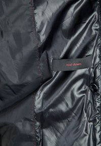 Sort, glat stof med en blank finish. Indeholder en rød stitched detalje og et mærke med teksten "ægte dun." Knaplukning er synlig.