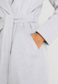 Grå, texturerad kimono med bälte i midjan, stora sidofickor och långa ärmar. Tyget verkar mjukt och varmt.