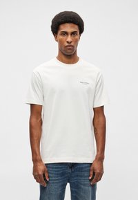 Λευκό βαμβακερό μπλουζάκι με κοντό μανίκι, κανονικό λαιμό και μικρό μαύρο λογότυπο στο αριστερό στήθος. Φοριέται με μπλε τζιν.