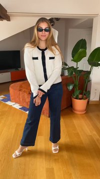 Baltas kardiganas su juodais akcentais, tamsiai mėlynos kelnės ir baltos suknelės bateliai su dirželiais. Modelis dėvi juodus ovalius akinius nuo saulės. Fonas su žaliąja augmenija.
