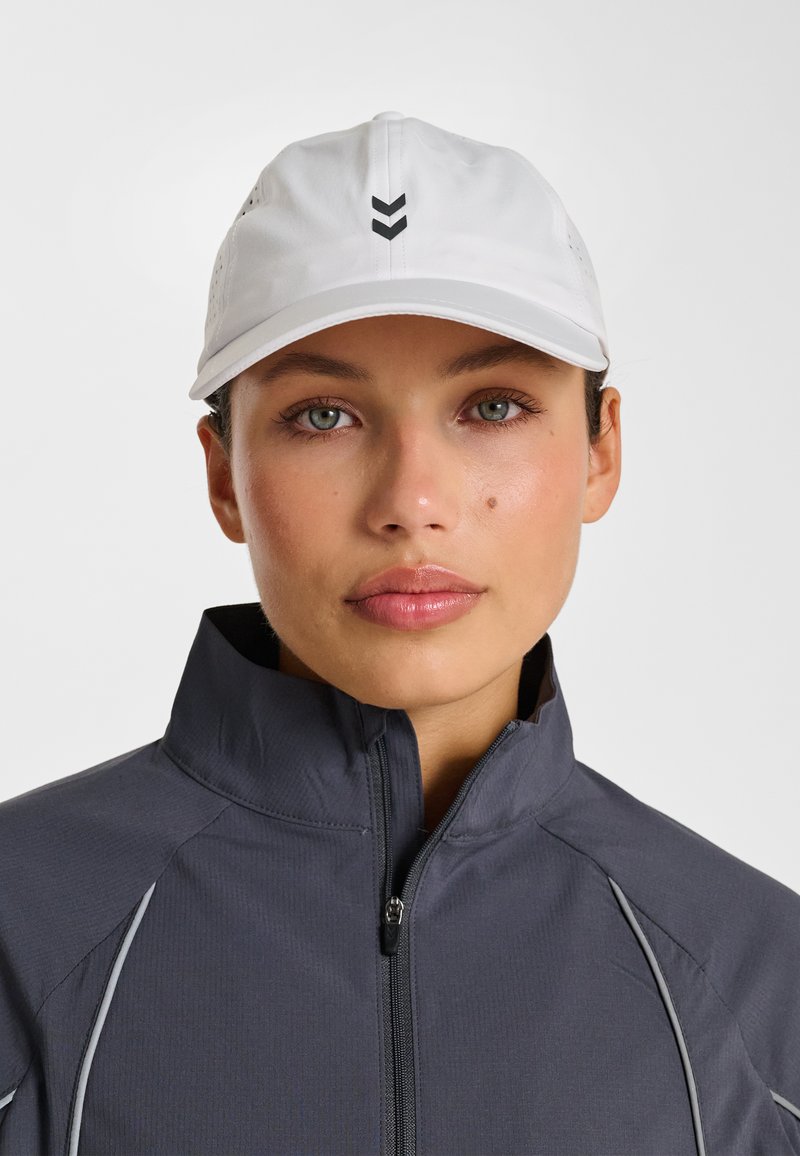 Vitt keps med ventilationshål, med en svart chevrondesign på framsidan. Bärs med en mörkgrå jacka med dragkedja.