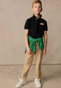 Černé polo tričko s límečkem, zelený svetr uvázaný v pase, khaki kalhoty a bílé tenisky. Na triku je vyšitý detail vlajky.