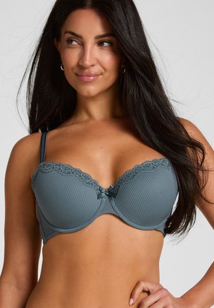 Donna con i capelli lunghi e scuri indossa un reggiseno blu con rifiniture in pizzo, guarda di lato con un leggero sorriso su uno sfondo chiaro e uniforme.