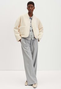 Giacca cropped in misto lana color crema con tasca frontale, camicia grigia con bottoni e pantaloni grigi larghi. Le scarpe presentano un design testurizzato color crema.