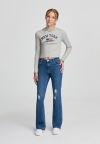 Felpa grigia crop con testo e grafica della bandiera americana. Jeans blu a campana con dettagli strappati. Sneakers bianche completano l'outfit.