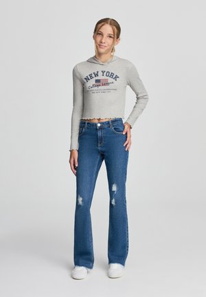 Felpa grigia crop con testo e grafica della bandiera americana. Jeans blu a campana con dettagli strappati. Sneakers bianche completano l'outfit.