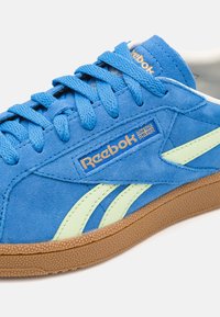 Sneaker blu scamosciato con dettagli verdi, suola in gomma marrone testurizzata e lacci blu. Presenta il logo Reebok in filo dorato sul lato.