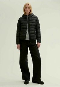 Casaco puffer acolchoado preto com capuz, fecho de correr e um suéter cinza por baixo. Modelo usa calças largas pretas e sapatos pretos robustos.