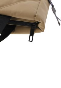 Esquina de una mochila de nylon beige con un zipper negro y un tirador. Textura suave con una forma estructurada y una correa negra en contraste.
