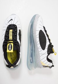 Nike Air Max-sneakers i vitt med svarta accenter, med ett rutmönster, transparent sula och en djärv gul etikett. Texturerad överdel.