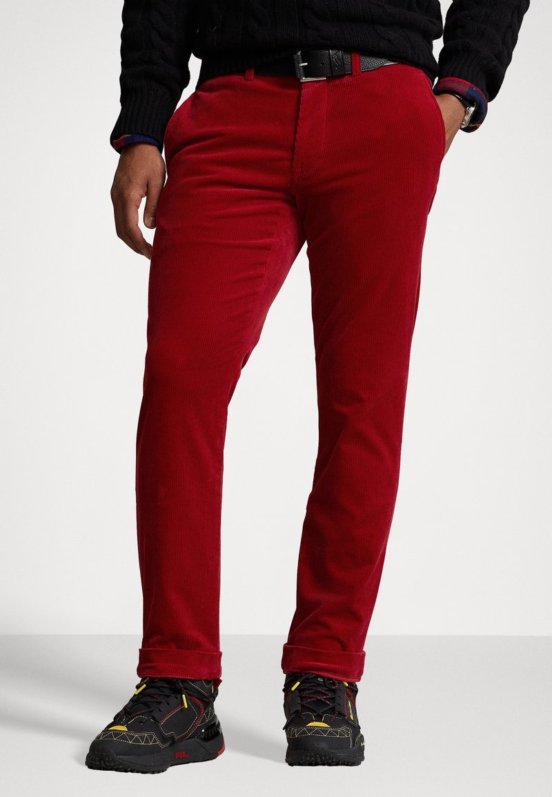 Mand iført rød slim-fit fløjlsbukser, sort bælte, sort strikket sweater og sorte sneakers med gule og røde detaljer, stående med hænderne i lommerne.