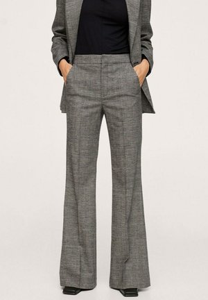 Vrouw draagt grijze geruite hoge taillebroek met uitlopende pijpen en bijpassend blazer over een zwart topje, handen in de zakken, rechtopstaand.