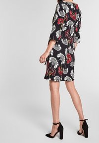Robe noire avec un motif floral rouge et blanc. Manches mi-longues et silhouette droite. Portée avec des chaussures à talons hauts noires.