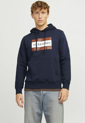 Sudadera azul marino de tejido suave que presenta un bolsillo frontal, capucha con cordón y un logo central en blanco y marrón sobre un parche rectangular.