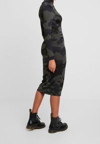 Långärmad, kroppsnära kamouflageklänning i mörkgröna och gråtoner, ihop med svarta snörstövlar och en glänsande yta.