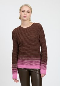 Jonge vrouw met kort blond haar, gekleed in een getextureerde bruine trui met roze manchetten en zoom, en donkerbruine leren broek, staande tegen een witte achtergrond.