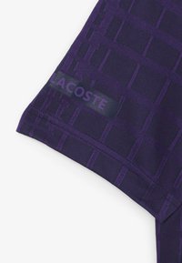 Lila idrottströja med rutmönster, slät textur och en Lacoste-logga närmast fållen.