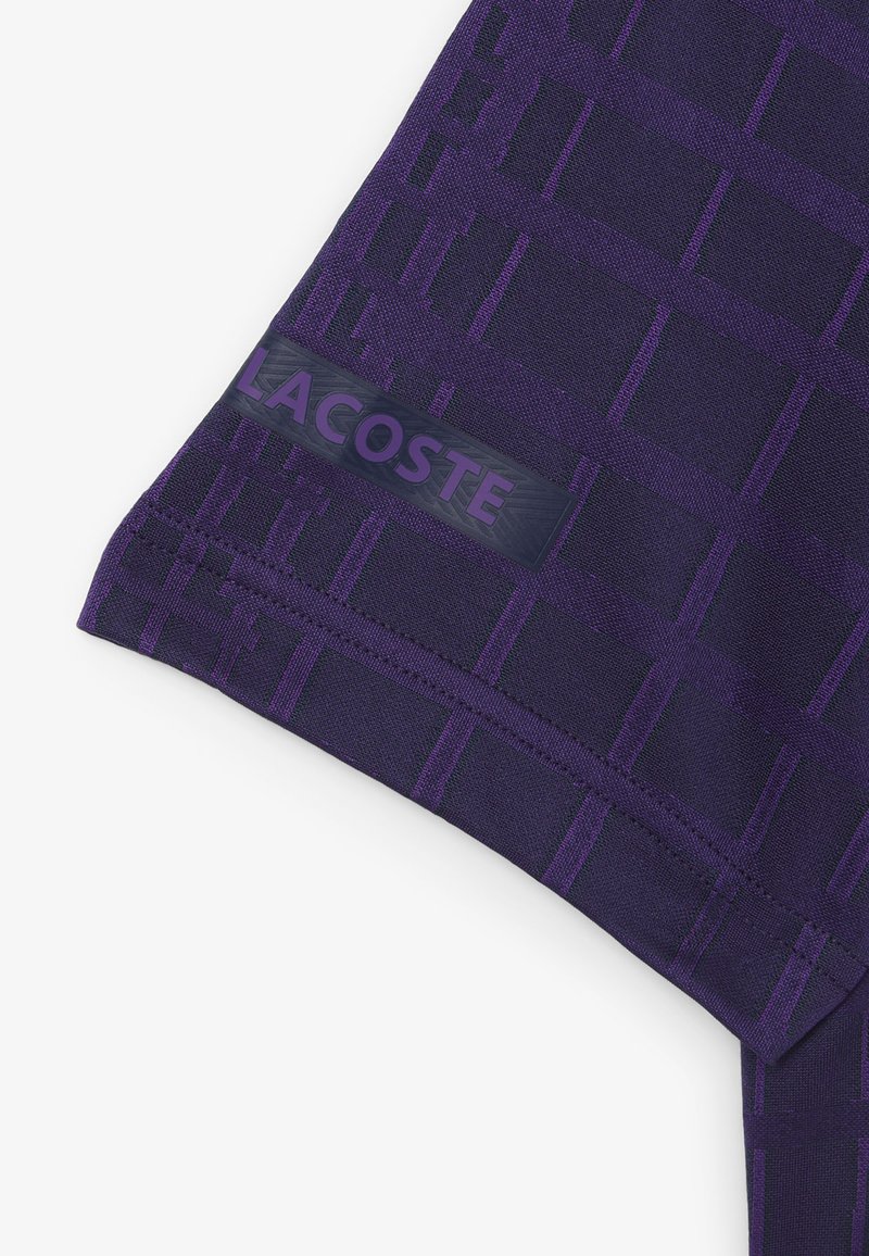Maglietta sportiva viola con motivo a griglia, texture liscia e un'etichetta con il logo Lacoste vicino all'orlo.