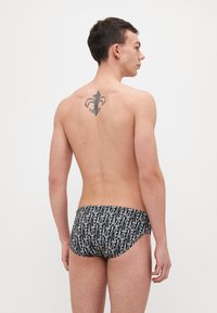 Slips de înot elastic în negru, cu text alb repetat. Prezintă o croială mică și o textură netedă, moale. Spate expus cu un tatuaj vizibil.