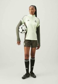Hele roheline lukuga sporditopp tume roheliste varrukatega, mustad lühikesed püksid ja mustad jalgpallisokid valgete triipudega. Mängija hoiab jalgpalli.