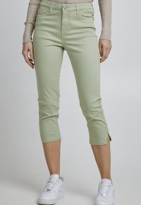 Jeans courts vert clair avec une coupe slim. Présente une taille haute, des fentes latérales à l'ourlet et une fermeture à bouton. Associés à des baskets blanches.