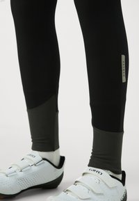 Czarne legginsy sportowe z kontrastującymi szarymi sekcjami na kostkach, w połączeniu z białymi butami rowerowymi z trzema paskami i teksturowanymi akcentami.
