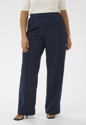 Donna con pantaloni ampi blu navy, top beige a maniche corte e sandali neri con tacco e punta quadrata, in piedi con le mani rilassate ai lati.