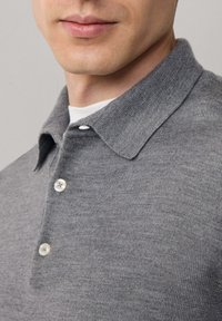 Grijze gebreide poloshirt met een kraag, met twee knopen en een zachte textuur, gedragen over een wit T-shirt, close-up van de halslijn.