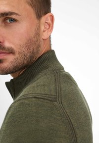 Nahaufnahme eines Mannes mit leichtem Stoppeln, der einen grünen Strickpullover mit einem hohen Rippkragen trägt und leicht zur Seite schaut.