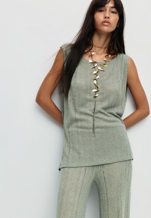 Haut sans manches vert clair avec texture en côtes verticales, assorti à un pantalon coordonné. Il est orné d'un collier décoratif en métal doré aux formes irrégulières.