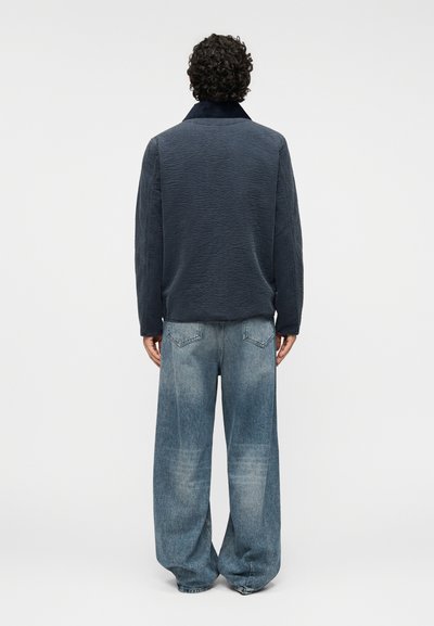 Ναυτικό μπλε μπουφάν με καπιτονέ, υψηλό γιακά, σε συνδυασμό με άνετο ανοιχτό μπλε τζιν παντελόνι. Το σύνολο έχει χαλαρή εφαρμογή και υφή υλικού.