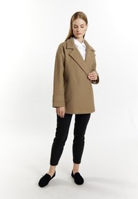 Beige wollenblend jas met brede revers, zijzakken en omgeslagen manchetten, gecombineerd met zwarte slim-fit trousers en donkere instap schoenen.