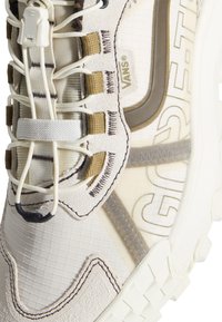 Gros plan d'une basket Vans beige avec des coutures texturées, des lacets à cordon et un tissu semi-transparent laissant apparaître un texte imprimé en dessous.