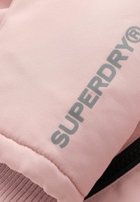 Chaqueta aislante de color rosa claro con textura cosida, un bolsillo con cremallera y el logo gris "SUPERDRY®". Tela suave con un acabado liso.
