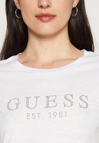Balta marškinėliai su žodžiu "GUESS", išsiuvinėtų blizgančiais sidabro spalvos akmenukais, apvaliu kaklu ir švelnia audinio tekstūra.
