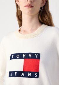 Hvit genser med ribbestrikket halslinning, preget av en dristig, vevd grafikk av "TOMMY" i marineblått, og "JEANS" i hvitt på en rød boks.