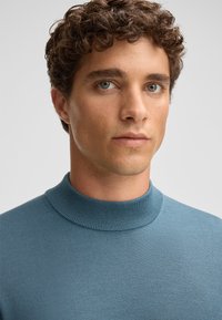 Helles bläuliches Strickpullover mit hochgeripptem Kragen und glatter Textur, auf einer Person vor einem neutralen Hintergrund präsentiert.