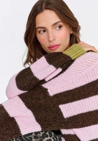 Sweter z dzianiny w dużych blokach brązu i jasnego różu, z zielonym akcentem przy mankiecie, ukazujący fakturę i luźny krój.