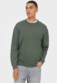 Groene gebreide sweatshirt met een ronde hals, geribbelde manchetten en onderrand. Model poseert met één hand in de zak, gecombineerd met lichtgrijze jeans.