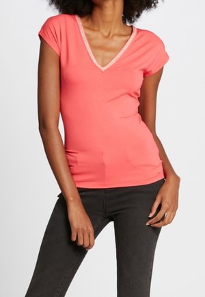 Femme portant un haut moulant corail à manches courtes et col en V, et un jean noir, debout avec une main près de la hanche et l'autre reposant sur sa cuisse.