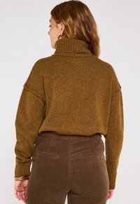 Maglione marrone lavorato a maglia con colletto alto e vestibilità ampia. Il tessuto testurizzato presenta delle macchie multicolori. Abbinato a pantaloni in velluto a coste marroni.